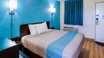 A hotel room with a bed, nightstand, wall-mounted lamp, air conditioning unit, and a window with curtains.
