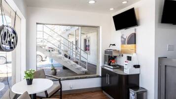 A modern hotel room with a large window, a flat-screen TV, a coffee machine, and a small table with a chair.