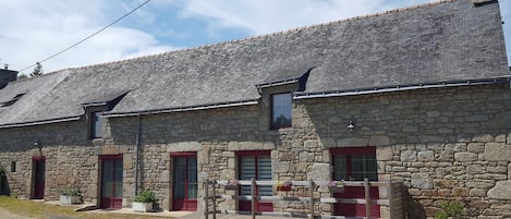 A stone building with a tiled roof, multiple windows with red frames, and a wooden fence in front.