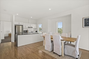 A modern kitchen with white cabinetry, a stainless steel refrigerator, and a wooden dining table with white chairs. - Coral Haven ~ Port Willunga - Seaside Sanctuary in a Prime Port Willunga Location (Port Willunga)