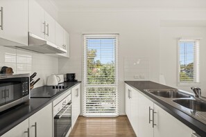 A modern kitchen with white cabinetry, a black countertop, a built-in oven, and a window with blinds. - Coral Haven ~ Port Willunga - Seaside Sanctuary in a Prime Port Willunga Location (Port Willunga)