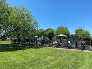 A playground with various play structures, a fenced area, and picnic tables. - KU Lake & Golf House (Pet Friendly) (Lawrence)