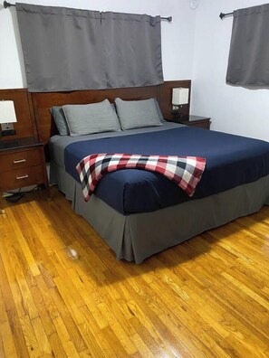 A bedroom with a wooden headboard, a bed with grey bedding, a checkered throw blanket, a nightstand with a lamp, and a wooden floor. - KU Lake & Golf House (Pet Friendly) (Lawrence)