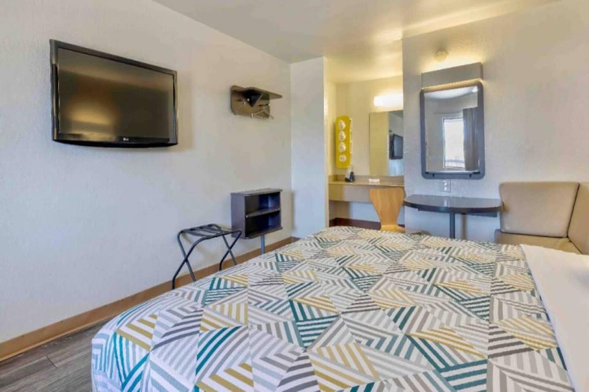 A hotel room with a bed, a television, a desk, a chair, a mirror, and a small shelf.