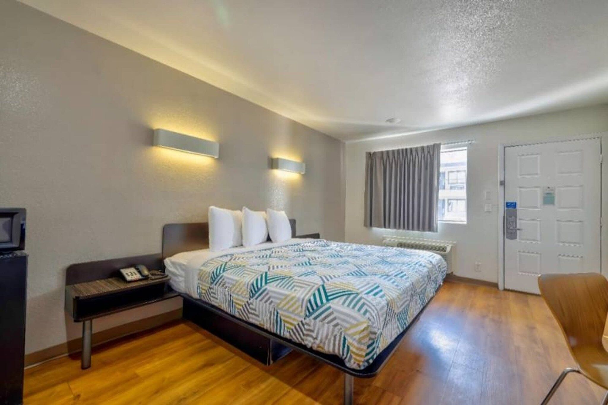 A hotel room with a bed, bedside table, lamp, window, and door.