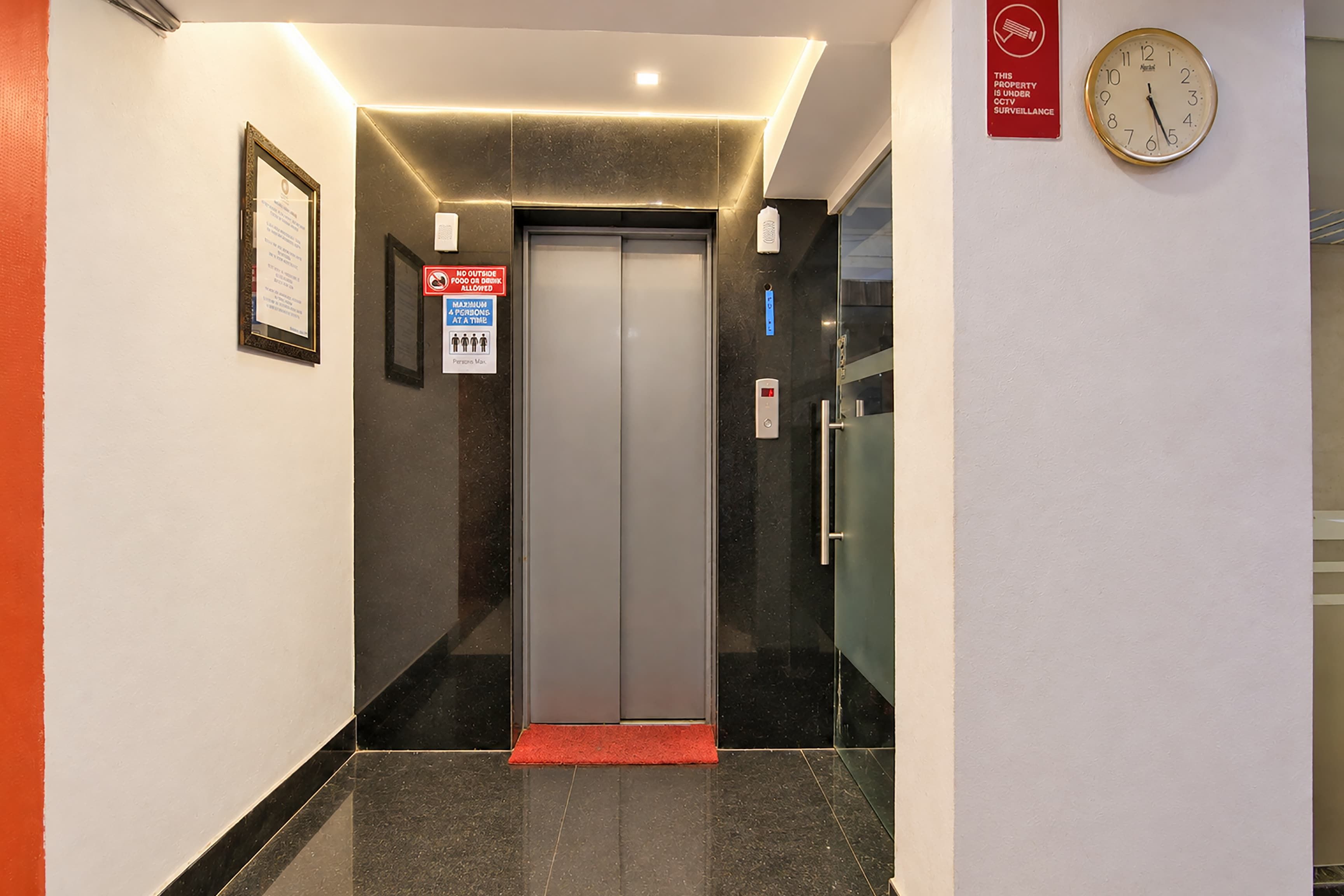 An elevator with a digital display, a red mat at the entrance, and a clock on the wall.