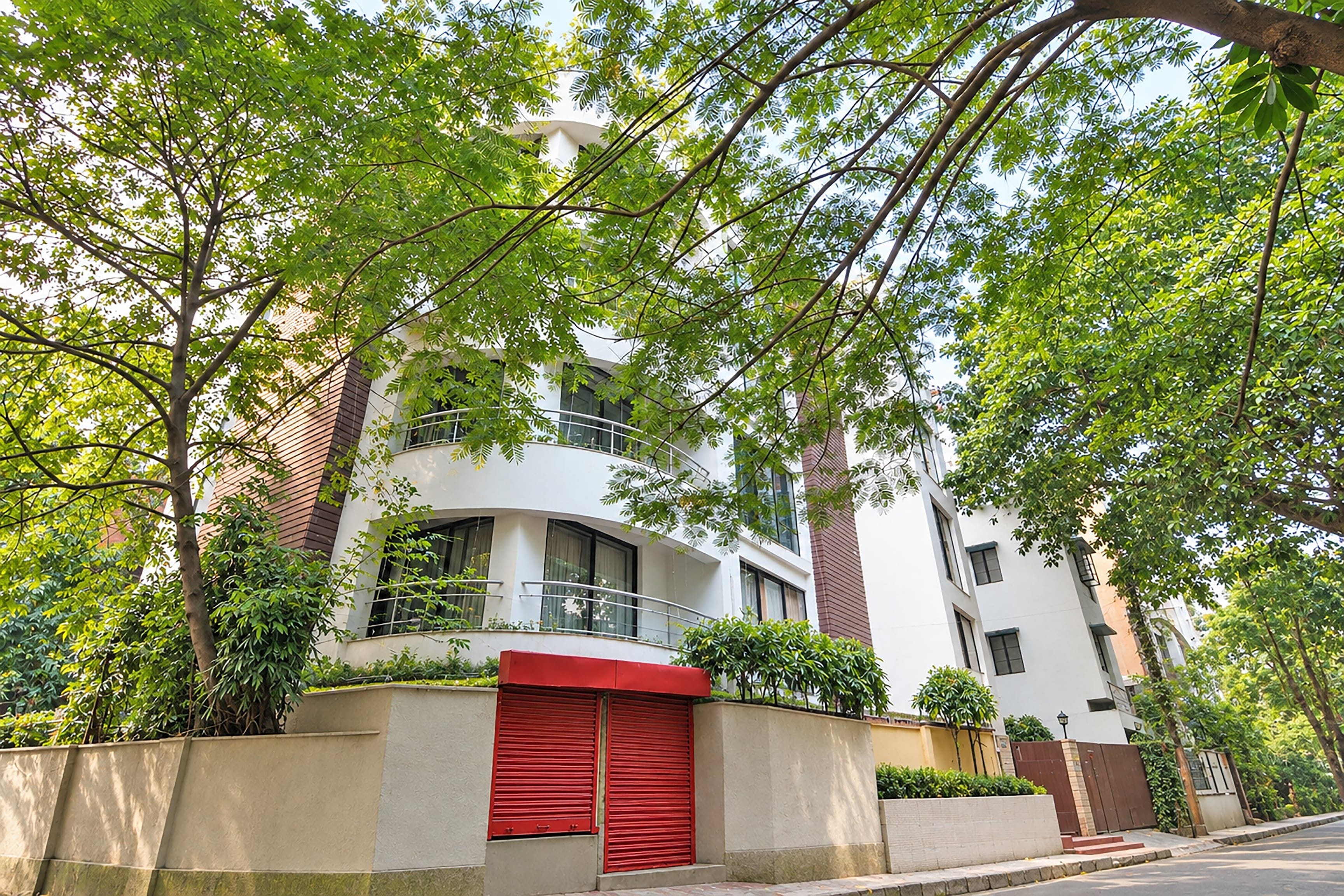 A modern multi-story residential building with a red door, surrounded by lush green trees.