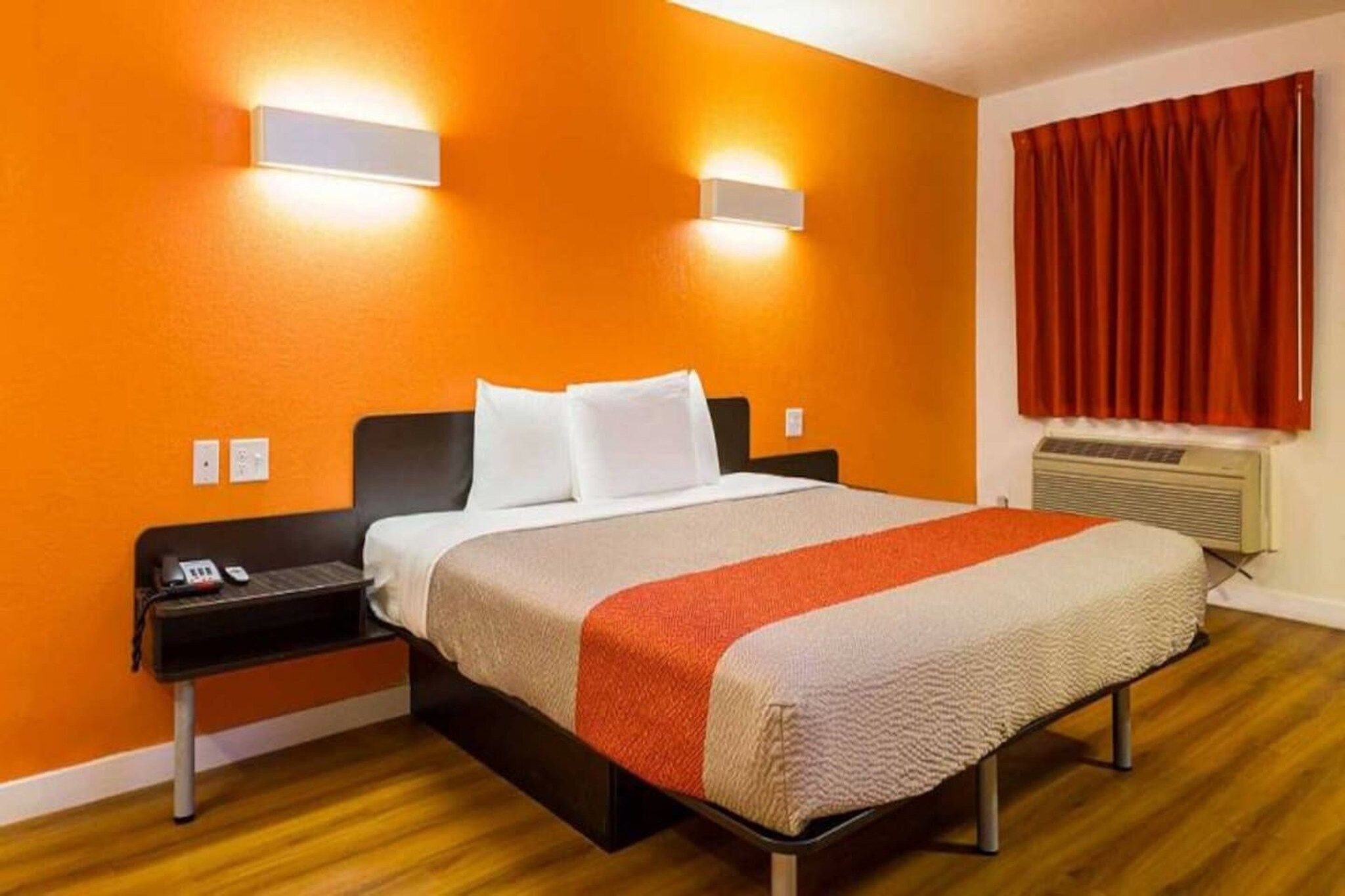 A hotel room with a bed, a nightstand, a wall-mounted lamp, a window with curtains, and an air conditioning unit.
