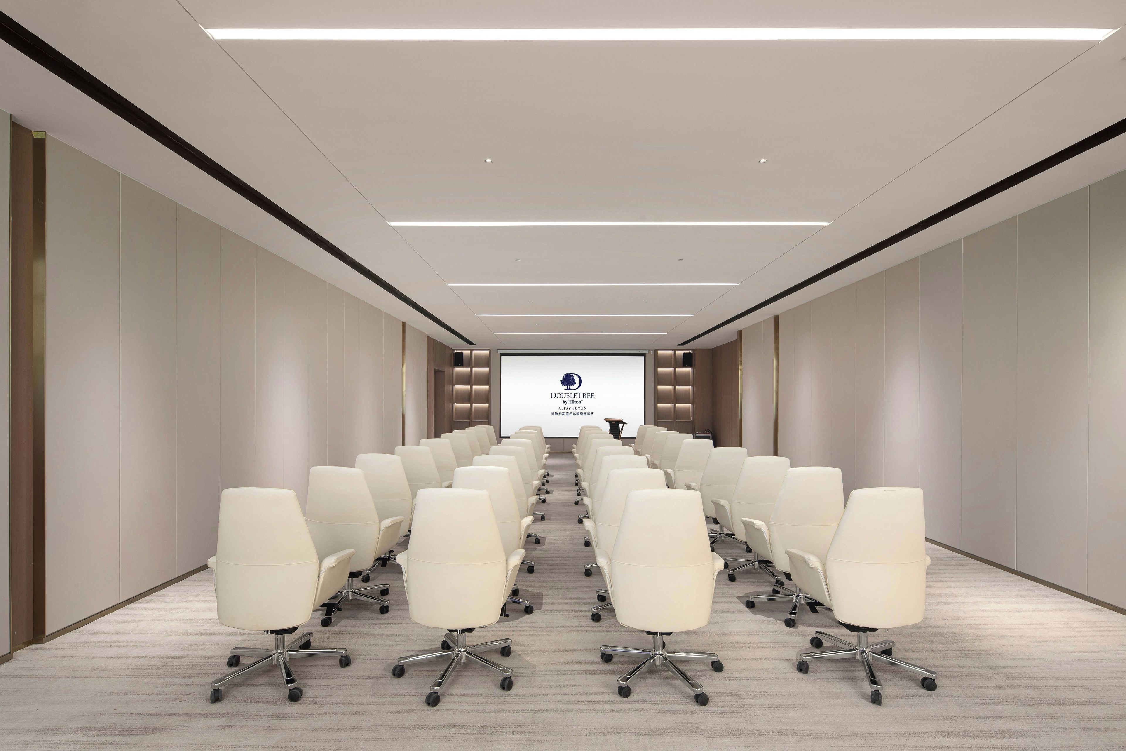Une salle de conférence avec des rangées de chaises blanches, un grand écran à l’avant et un design moderne au niveau du plafond.