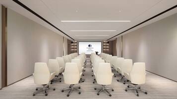Une salle de conférence avec des rangées de chaises blanches, un grand écran à l’avant et un design moderne au niveau du plafond.
