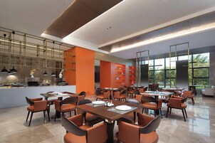 A modern dining area with multiple tables and chairs, a kitchen in the background, and large windows providing natural light.