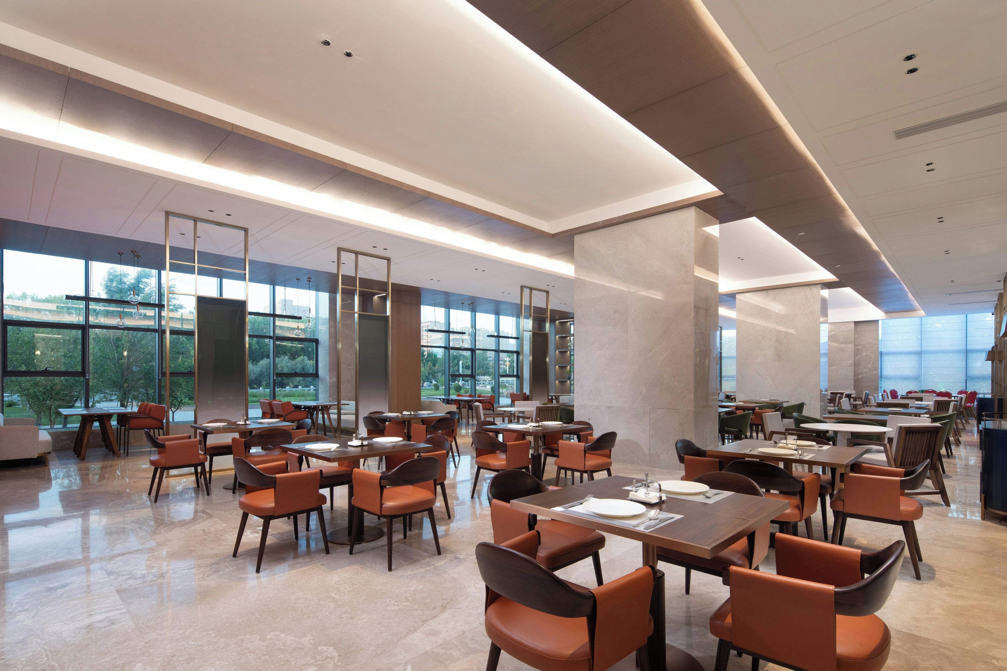 Un'ampia sala da pranzo con diversi tavoli e sedie, grandi finestre che offrono vista sull'esterno e un design architettonico moderno caratterizzato da linee pulite e colori neutri.