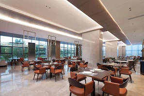 Un comedor espacioso con varias mesas y sillas, ventanas amplias que ofrecen vistas al exterior, y un diseño arquitectónico moderno con líneas limpias y una paleta de colores neutros.