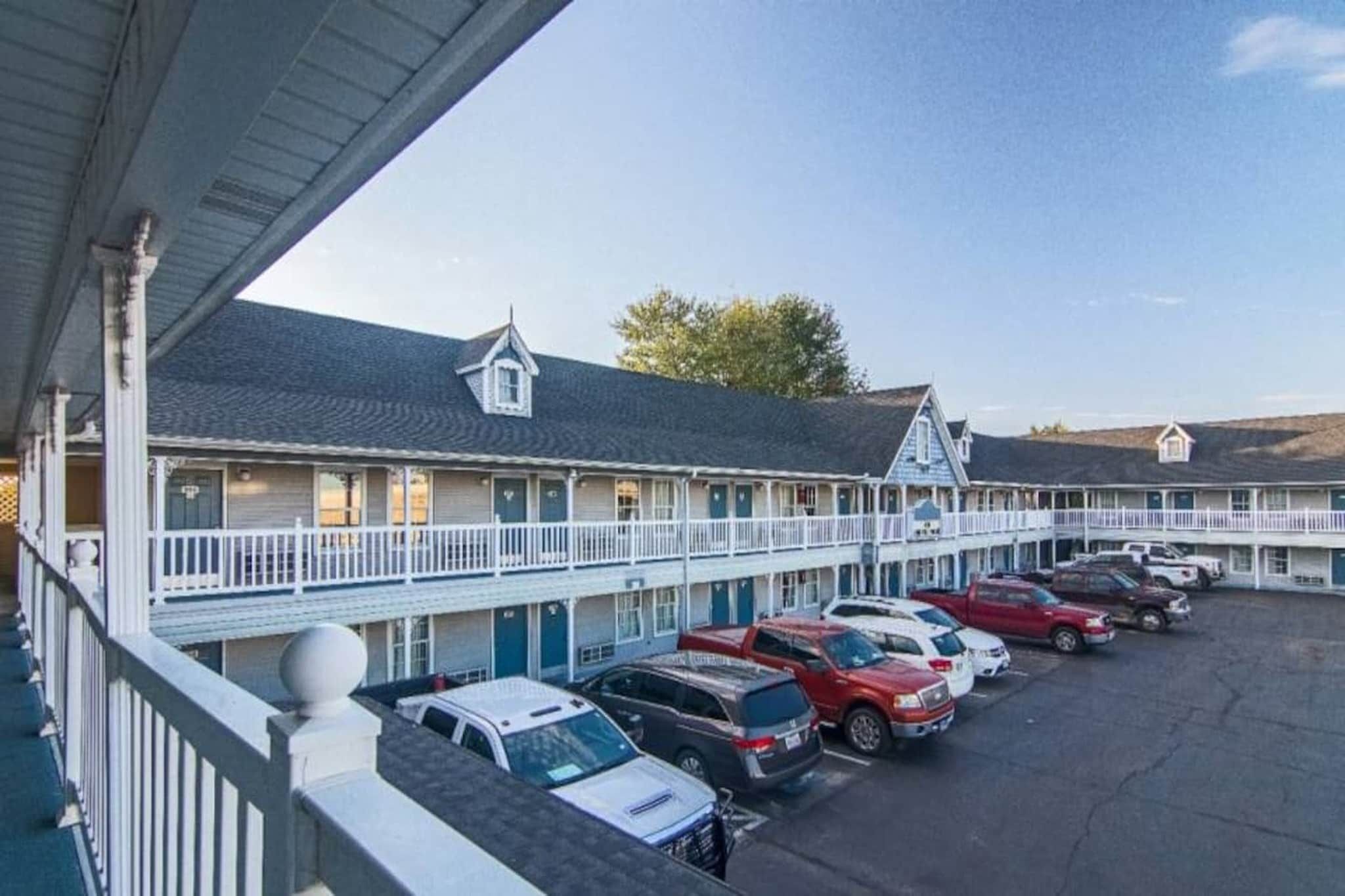 Un motel avec une rangée de chambres, chacune dotée d’un balcon et d’un petit porche. Un parking se trouve devant, où plusieurs voitures sont garées.