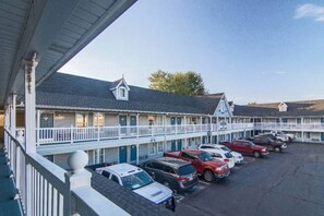 Un motel con una hilera de habitaciones, cada una con un balcón y un pequeño porche. Hay un estacionamiento con varios coches aparcados al frente.