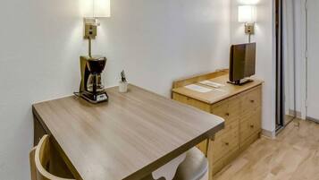 A small, simple room with a wooden table, a coffee maker, a television on a wooden cabinet, and a door.