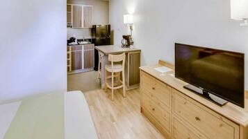 A compact hotel room with a bed, a TV, a small desk with a chair, a kitchenette, and a wooden cabinet.