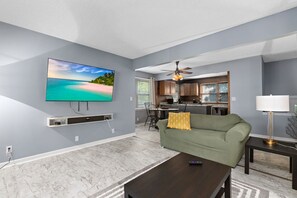 A living room with a flat-screen TV, a sofa, a coffee table, and a kitchen in the background. - Charming 4br house w/ Free parking + Wifi (De Soto)