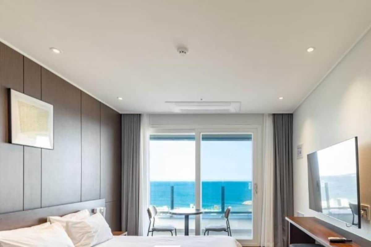 A hotel room with a bed, a desk, a chair, a TV, and a large window with a view of the ocean.