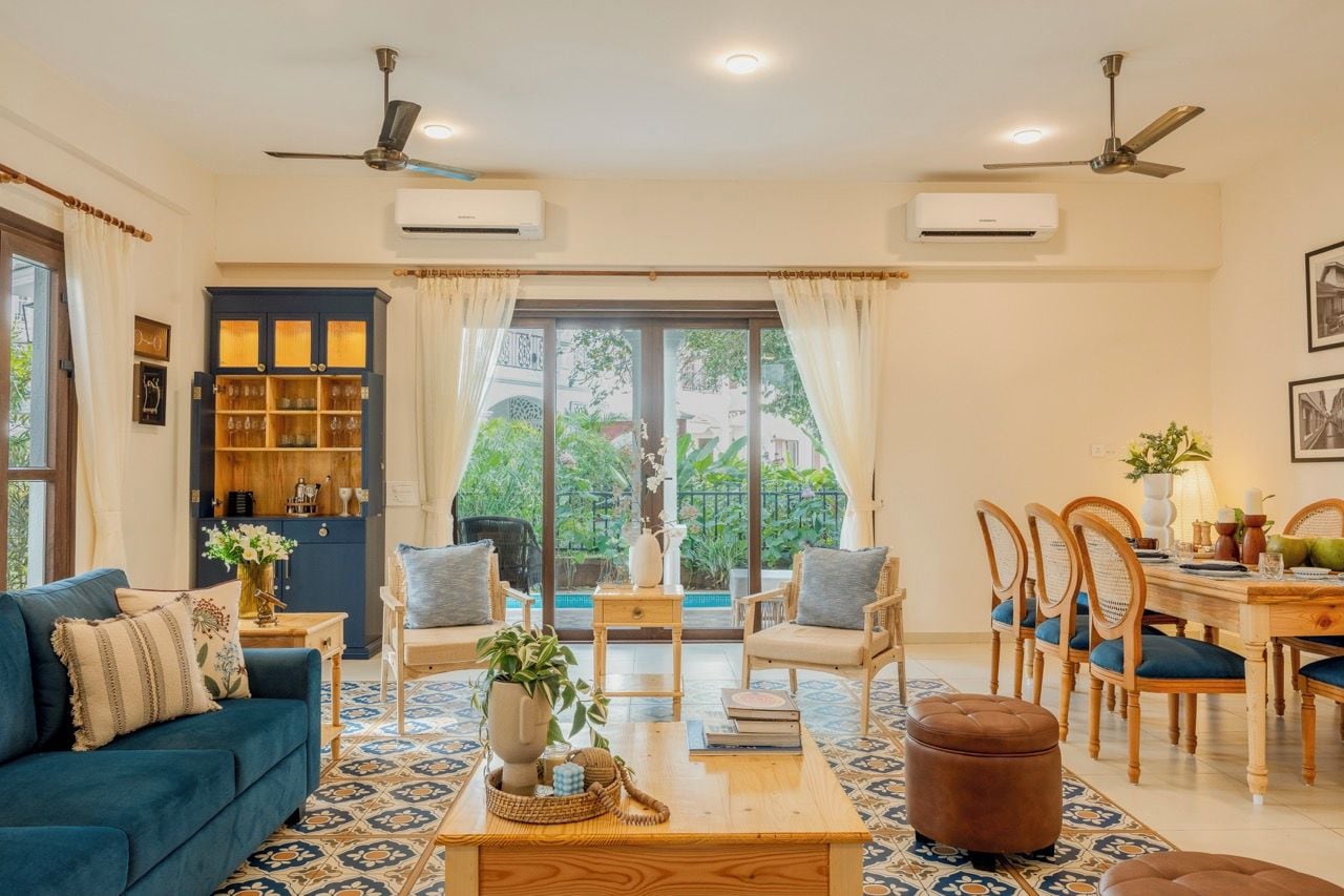A living room with a sofa, coffee table, and dining area with chairs. There are ceiling fans, an air conditioning unit, and a view through large windows.