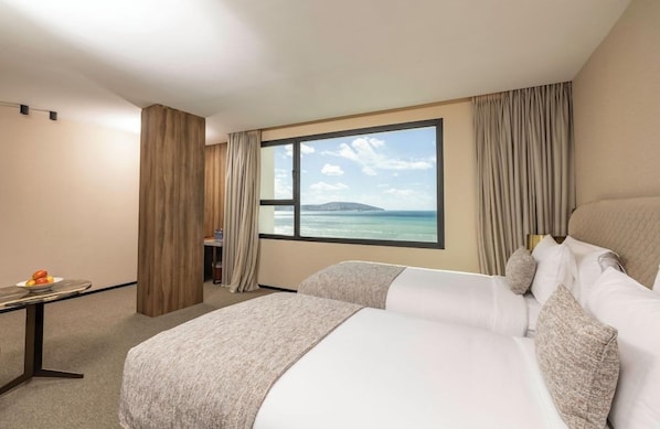 Habitació d'hotel amb una finestra gran, un llit, una taula amb fruita i vista al mar.
