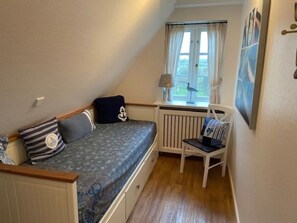 A small, cozy room with a sloped ceiling, a single bed with patterned bedding, a chair with cushions, a window with curtains, and a radiator. - Deichhof Holiday Apartment 8 (Dunsum)