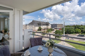 A balcony with a table and chairs, a vase with flowers, and a view of buildings and a street. - Holiday apartment with 2 bedrooms (Westerland - Sylt)