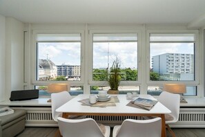 A dining area with a table set for four, large windows with blinds, and a view of buildings and greenery outside. - Holiday apartment with 2 bedrooms (Westerland - Sylt)