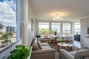 A modern living room with a sofa, dining table, and chairs, large windows with a city view, and a ceiling light fixture. - Holiday apartment with 2 bedrooms (Westerland - Sylt)