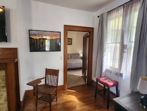 Ein Zimmer mit Holzboden, einem Holzbstuhl, einer roten Bank, einem Bett im Hintergrund und einem Gemälde an der Wand.