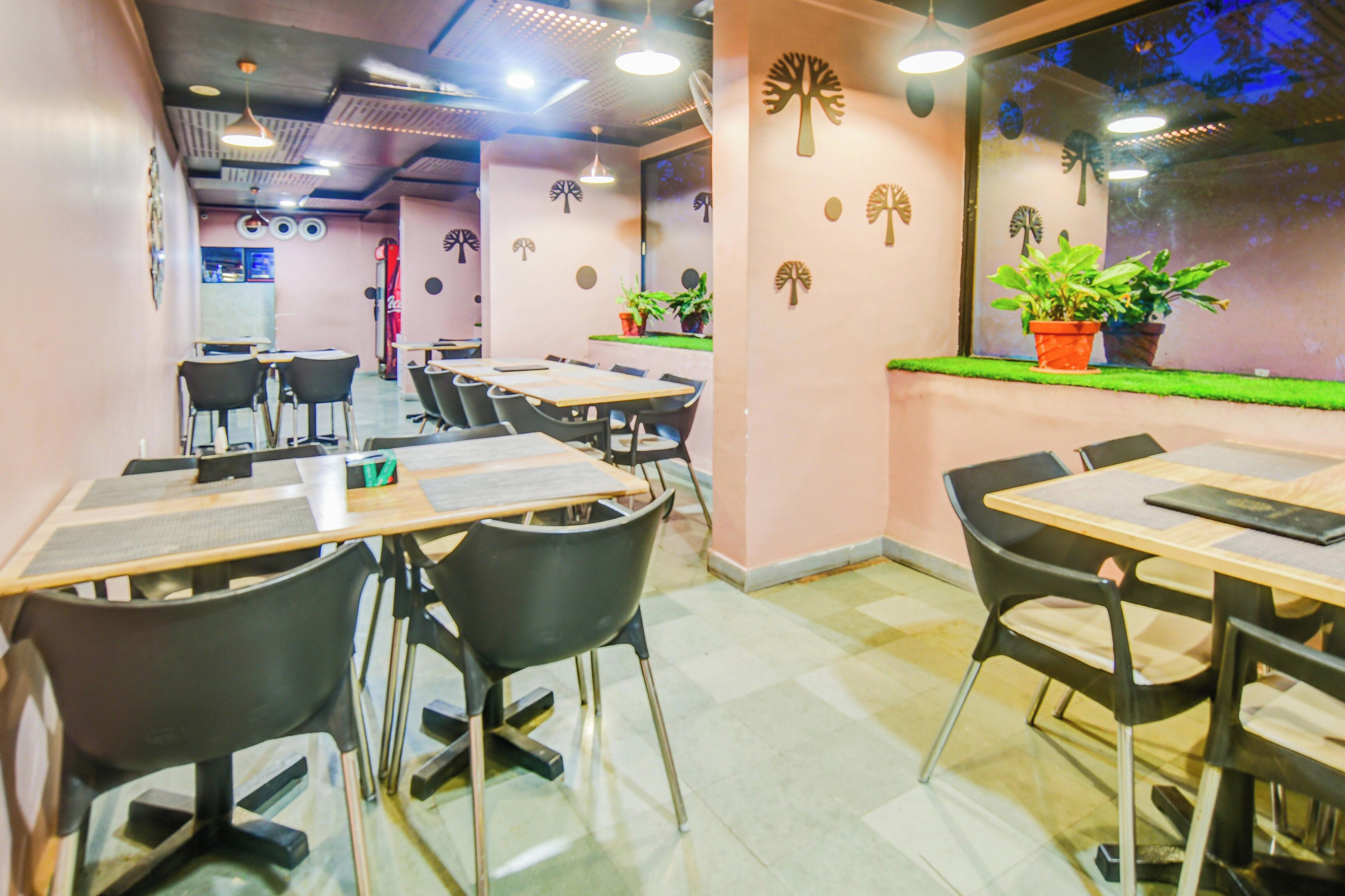 A dining area with tables and chairs, a wall with tree decorations, and a window with plants.