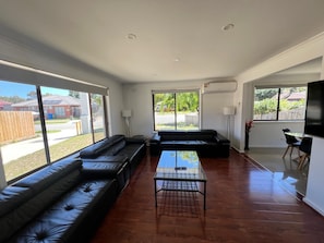 A modern living room with a black leather sofa, a glass coffee table, and large windows providing a view of the outside. - Beautiful and spacious house in Doveton (Doveton)