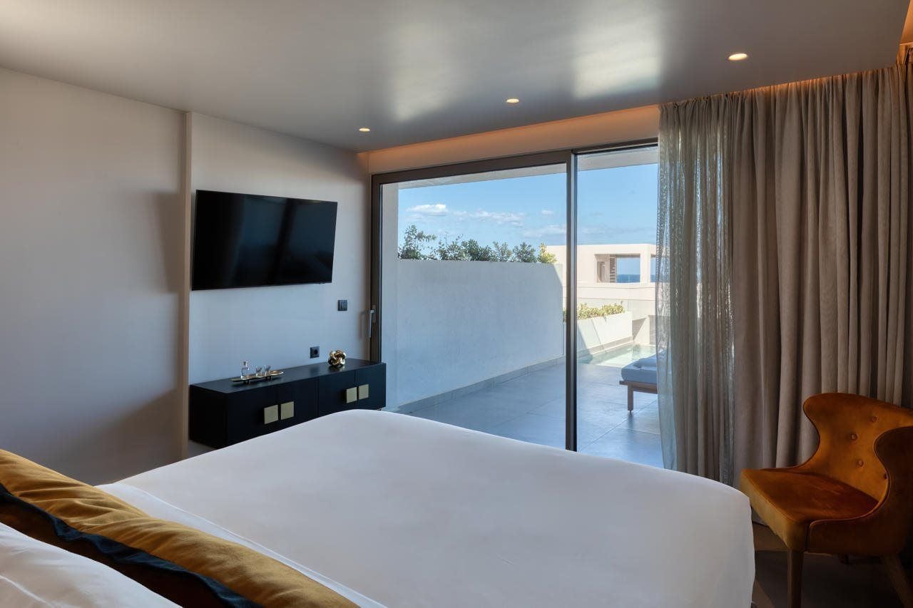 Una habitación de hotel moderna con una cama grande, un televisor de pantalla plana montado en la pared, una mesita de noche pequeña y una puerta corredera de vidrio que da a un espacio exterior.
