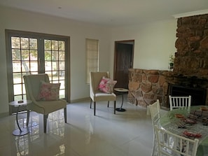 A spacious room with a stone fireplace, two chairs with floral cushions, a dining table with a glass top, and a large window with blinds.