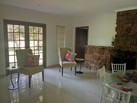 Una habitación amplia con chimenea de piedra, dos sillones con cojines estampados, una mesa de comedor con cubierta de vidrio y un gran ventanal con persianas.