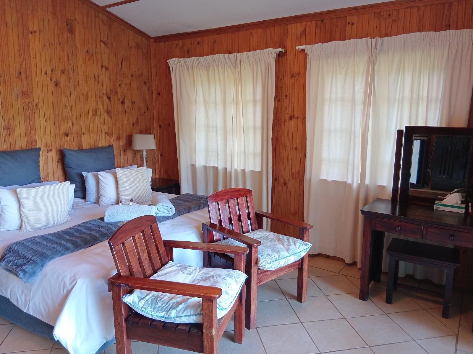 A hotel room with a wooden paneled wall, a bed with multiple pillows, two wooden chairs with cushions, a desk with a mirror, and curtains.