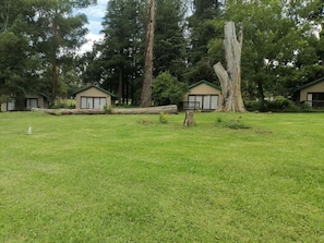 A grassy area with two buildings, each with a large window and a door, surrounded by trees.