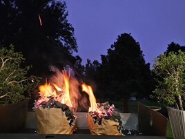 Escena nocturna al aire libre con un fuego ardiendo en un contenedor metálico, rodeado de árboles.