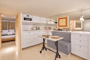 Une chambre d’hôtel compacte dotée d’une kitchenette, d’un coin repas avec un banc à carreaux, d’un lit avec du linge de lit blanc et d’un grand miroir.