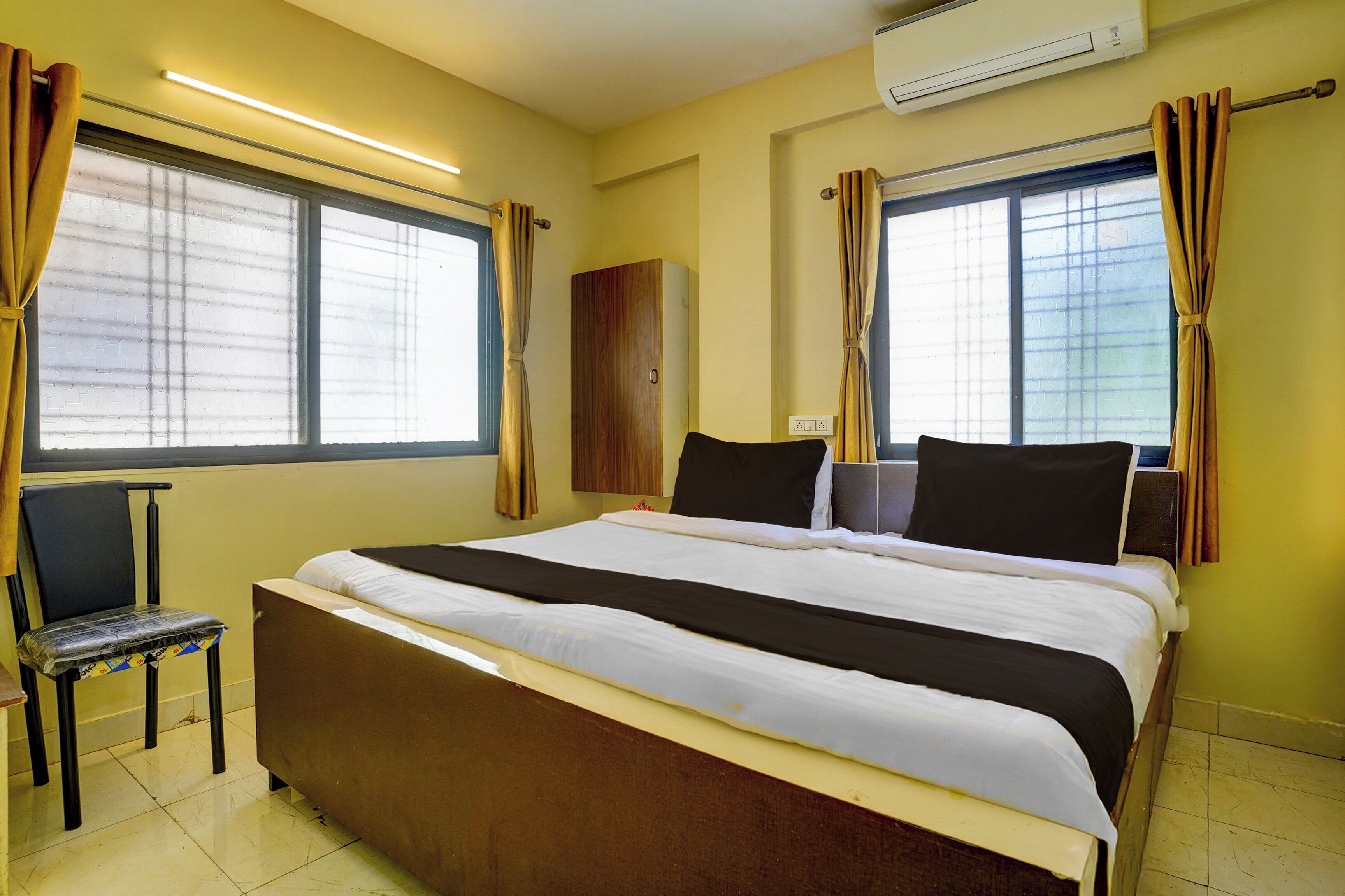 Habitación de hotel con una cama grande, dos ventanas con cortinas, una silla, un armario y un equipo de aire acondicionado.