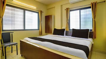 Habitación de hotel con una cama grande, dos ventanas con cortinas, una silla, un armario y un equipo de aire acondicionado.