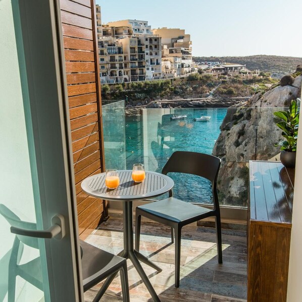 Un balcón con mesa y sillas de vidrio, con vistas a una zona costera con edificios y un paisaje rocoso.