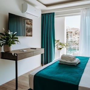 Habitación de hotel con cama, escritorio, planta en maceta y vista a edificios a través del ventanal.