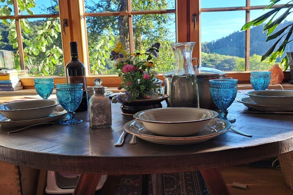 Mesa de madera redonda con una botella, jarra y cristalería azul, frente a una ventana que da a vistas de vegetación.