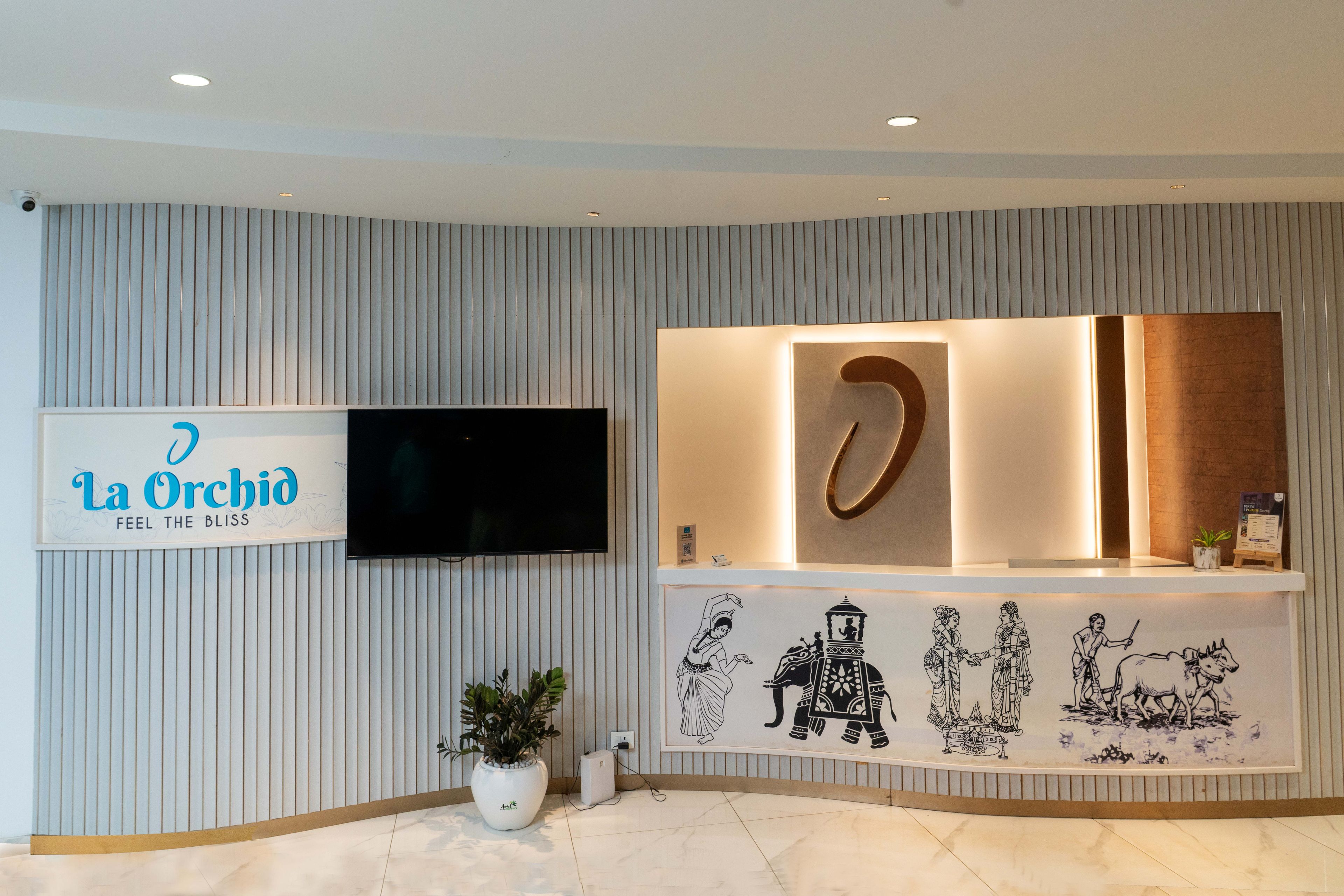 Una zona de recepció amb un senyal que diu 'La Orchid FEEL THE BLISS', un televisor de pantalla plana, una paret decorativa amb obra d'art cultural i una planta en test.