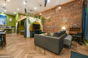 A modern living room with a herringbone wooden floor, a grey sofa, a brick wall, and a television mounted on a wooden arch. - Artsy studio in Lawrenceville: easy & free parking (Pittsburgh)