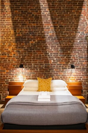 A bed with a headboard, a bedside table, and a wall with a brick pattern. - Artsy studio in Lawrenceville: easy & free parking (Pittsburgh)