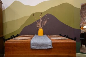 A dining table with a patterned runner, a yellow vase with dried plants, and a mural of a mountain landscape in the background. - Artsy studio in Lawrenceville: easy & free parking (Pittsburgh)