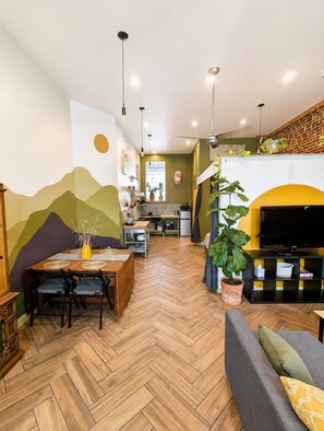A modern living room with a herringbone floor, a dining area with a wooden table, and a TV on a black stand. - Artsy studio in Lawrenceville: easy & free parking (Pittsburgh)