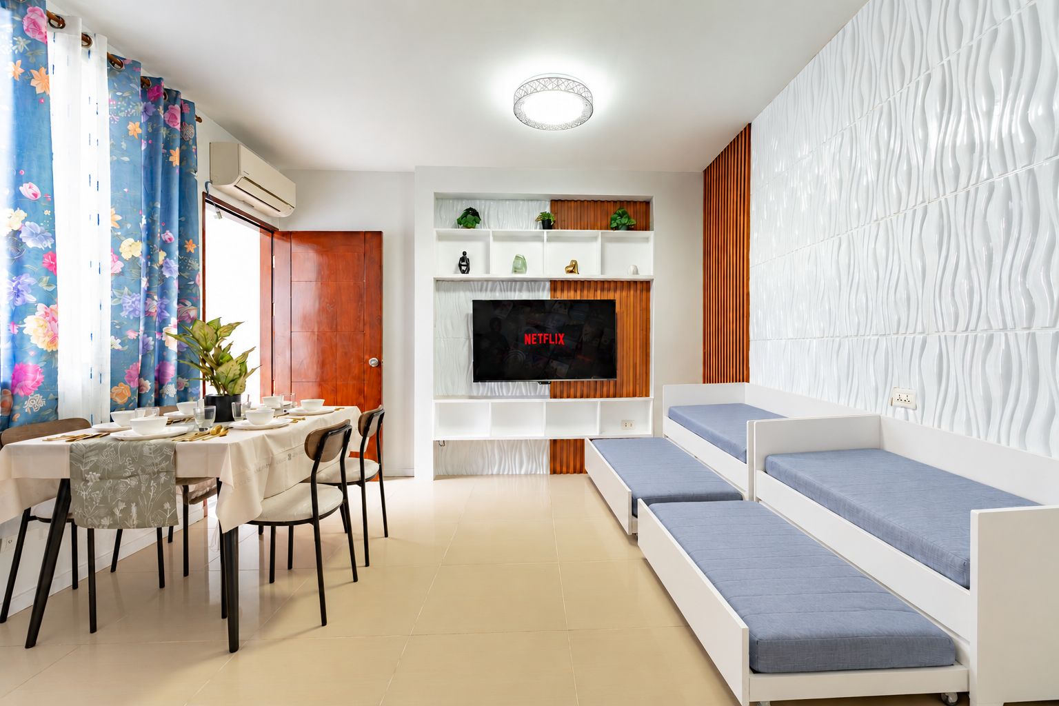 Een moderne woonkamer met een eetgedeelte, een flatscreen televisie aan een houten wand en een bank met blauwe kussens.